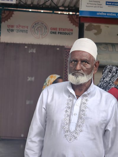 The image shows an elderly man with dark skin, a white beard, and a white skullcap, wearing a white kurta with intricate embroidery around the neckline. He is looking directly at the camera with a serious expression. In the background, there is a sign in Hindi that reads "एक स्टेशन एक उत्पाद" (Ek Station Ek Utpad), which translates to "One Station One Product". Another part of the sign, in English, reads "ONE STATION ONE PRODUCT". Behind him, there are other people partially visible and a sign for "Bank of India". The overall setting appears to be an outdoor public space, possibly a market or a train station, given the signage. The man's attire and the presence of the "One Station One Product" initiative suggest a context related to local commerce or government schemes. The details of the sign are clear enough to confirm it refers to a product available at a particular station. The environment is somewhat blurred in the background, focusing attention on the man. The lighting suggests it is daytime.