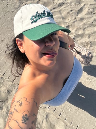 The image shows a close-up, high-angle selfie of a person wearing a white and green baseball cap, with the brim casting a shadow over their eyes. The person is also wearing a light blue halter top. Their tongue is sticking out slightly. On their left arm, there is a tattoo with several distinct designs, including a circular symbol with lines radiating outwards and a figure with spiky hair. The person is lying on a sandy beach, with the sand showing footprints and some small wooden stakes in the foreground. A rolled-up towel is visible behind the person's shoulder. The lighting suggests it is daytime and sunny. The image was taken on the sandy beach of Castelló de la Plana, Spain.