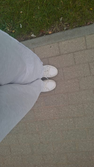 A first-person perspective shot from above shows a person's legs and feet, clad in light grey pants and white sneakers, standing on a brick pathway. The pathway is bordered by a strip of green grass. The scene is set in the city of Stowbtsy, Belarus. The lighting suggests it is daytime, likely overcast due to the soft shadows.