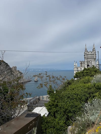 The image shows a sideways view of Swallow's Nest castle in Гаспринский поселковый совет, Ukraine, situated on a cliff overlooking the Black Sea. The castle, a prominent white structure with turrets, is partially obscured by green vegetation. Below the castle, the sea extends towards the horizon. In the foreground, there are rocky cliffs and foliage. A power line cuts across the view. Buildings and structures are visible on the cliffside.