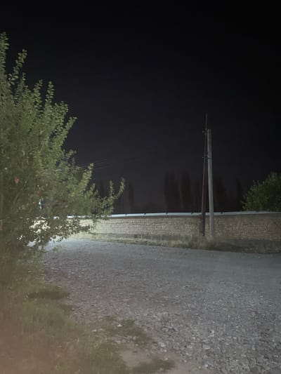 The image depicts an outdoor scene at night, likely in the city of Кедей-Арык, Kyrgyzstan. In the foreground, a dirt or gravel road is visible, bordered by sparse grass and a leafy tree on the left. In the middle ground, a low, brick wall stretches horizontally across the frame. To the right of the wall, a wooden utility pole stands, with electrical wires extending from it. In the background, silhouetted trees are visible against a dark night sky, with a faint hint of distant lights. The overall impression is of a rural or semi-urban setting at night, possibly on the outskirts of a settlement. There are no people or animals visible in the image.