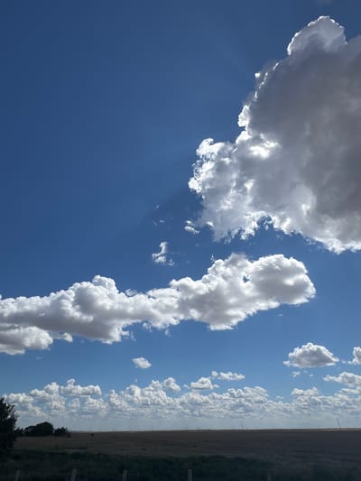 A wide, low-angle shot captures a vast expanse of blue sky filled with scattered white clouds. The clouds vary in size and density, with some appearing fluffy and bright, while others are darker and more substantial, partially obscuring the sun. Rays of light emanate from behind a large cloud in the upper right quadrant, creating a dramatic visual effect. The horizon line is visible in the lower portion of the image, separating the sky from a flat, brown, agricultural landscape. A faint line of trees and distant structures, possibly including wind turbines, are discernible on the horizon. The foreground features a dark, out-of-focus band of vegetation and what appear to be fence posts, suggesting the image was taken from a roadside or field's edge. The scene evokes a sense of open space and a clear, possibly windy, day in a rural setting. The location is identified as Shiderti, Kazakhstan. There are no people or animals visible in the image.