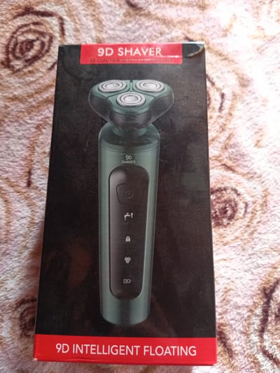 The image shows the box of a 9D SHAver. The box is predominantly black with a red band at the top and bottom. The top red band has white text that reads "9D SHAVER" and "9D CONTOUR DETECT TECHNOLOGY". The bottom red band has white text that reads "9D INTELLIGENT FLOATING". A green electric shaver with three rotating heads is depicted on the black portion of the box. The box is placed on a textured surface that appears to be a fabric with a swirled pattern. There are no people or any contextual information about the city of Monastirische, Russia visible in the image.