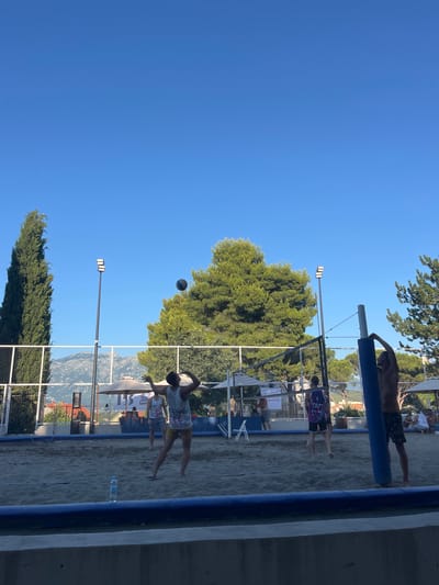 The image depicts a beach volleyball game taking place on a sandy court, with a clear blue sky overhead. In the foreground, a person wearing dark patterned shorts is standing next to a blue volleyball net post, with their hands on the post. To the left, a person in a white tank top and yellow shorts is in the process of hitting a volleyball, which is suspended in the air above them. Another person in a white tank top and dark shorts is in the background, near the net. A bottle of water is placed on the sand near the foreground. The court is bordered by a blue inflatable barrier. In the midground, there are beach umbrellas, trees, and mountains in the distance. Lights on poles are visible on the left and right sides of the court. The overall setting suggests an outdoor recreational area, possibly in Gošići, Montenegro, given the surrounding natural elements.