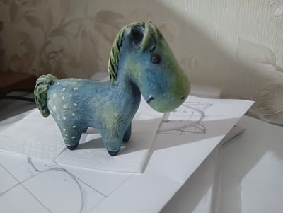 A close-up, slightly angled, eye-level shot captures a handmade, blue and green toy horse with white dots on its body, positioned on top of a white paper napkin. The horse is facing right, with its head turned slightly towards the viewer, revealing a dark eye and a black muzzle. The horse's mane is depicted with green strands. Behind the napkin, a sheet of white paper with faint pencil sketches of circles and lines is visible, casting a shadow from the horse. The background features a textured, off-white wallpaper with a floral pattern. The scene appears to be indoors, on a flat surface, possibly a desk or table. The lighting suggests a room with ambient light and possibly a nearby window creating a soft shadow on the wallpaper. The image does not provide information to determine a specific location in Bender, Moldova.