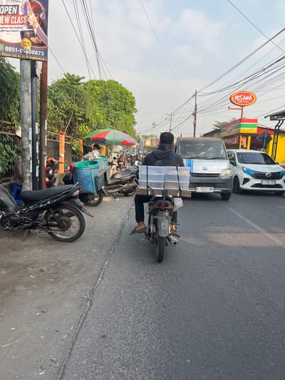 The image depicts a street scene in Kedungkendo, Indonesia, on a partly cloudy day. A person is riding a motorcycle, carrying several clear plastic containers filled with food items on a wooden rack attached to the back. The motorcycle is in motion on an asphalt road. To the left of the road, a sidewalk is lined with parked motorcycles, a blue plastic bin, and a multi-colored umbrella. Further left, a pole displays advertisements. On the right side of the road, a white car and a silver van are also in motion. In the background, a restaurant with a prominent red and yellow sign that reads "HISANA FRIED CHICKEN" is visible, along with other buildings and power lines. The overall environment appears to be a busy urban or suburban area.