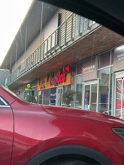The image displays a two-story commercial building facade at the Dockside Outlet Centre, Maritime Way, Chatham, Kent, United Kingdom. The photograph is taken from the interior of a red vehicle, with its glossy hood and front fender occupying the foreground. The building's ground level features several retail and leisure establishments. From left to right, partially visible storefronts include a "Subway" restaurant sign. Next is "Nando's," indicated by its illuminated red text sign and a circular red rooster logo, with two small, potted olive trees flanking its glass entrance doors. Adjacent to Nando's is "Gymfinity Kids Clubs & Nurseries." Further right, another storefront exhibits a partial Union Jack graphic on its facade. The upper level of the building features a long, metal-railed balcony with a corrugated metal facade above it. A large sign for "NINJA WARRIOR" is prominent on the upper level, positioned above the "Gymfinity Kids" and Union Jack storefronts. The scene is observed under clear daylight conditions. No individuals are discernible.