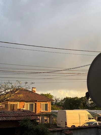 The image depicts a scene in Manole, Bulgaria. A two-story brick building with windows is partially visible on the right. Adjacent to the building is a white van. Power lines stretch across the sky, which is divided into areas of gray clouds and golden light. In the foreground, a gray, round object partially obscures the view. Vegetation is also visible on the right side of the image.