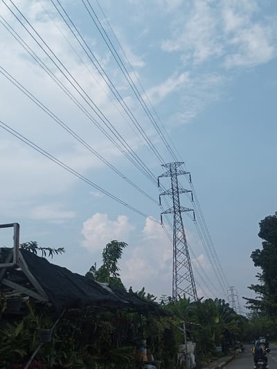 The image captures a daytime scene dominated by a large electrical transmission tower against a bright sky. Multiple parallel power lines stretch diagonally from the upper left to the upper right across the frame, converging towards the tall metal lattice tower, which stands prominently in the mid-right. The sky is a clear, light blue with scattered white, fluffy clouds.
In the foreground on the left, a structure covered with a dark, possibly black, tarp or shade netting is visible, likely a canopy or roof of a roadside stall. Beneath and around this structure, numerous green plants and foliage are densely packed, some appearing to be in pots, suggesting a plant nursery or a vendor selling plants. Part of a light-colored wooden or metal frame of this structure is visible on the far left.
To the right of the plant stall, more dense green vegetation, including trees and bushes, lines what appears to be a paved road. The road curves slightly into the distance on the right side of the image. On the road, a few vehicles, including what looks like motorcycles with riders, are visible in the background, moving away from the viewer. In the far distance, along the line of power lines, another smaller, similar electrical tower can be seen, indicating the extent of the power grid.
The lighting suggests it is bright daylight, possibly midday or early afternoon, given the intensity of the light and the appearance of the sky. The overall scene depicts a blend of natural elements (sky, clouds, lush greenery) and human infrastructure (power lines, towers, road, stall). There is no visible text in the image.