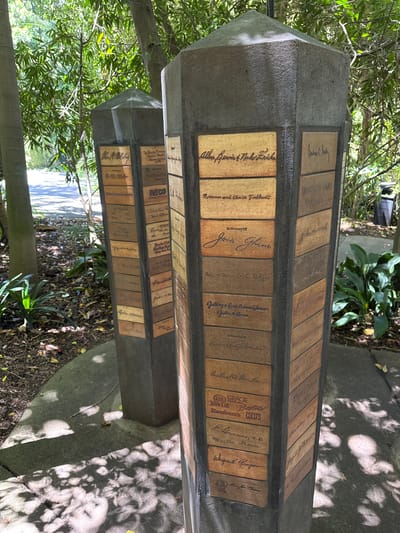 The image shows two memorial pillars in an outdoor, shaded area, likely a park or garden. The pillars are hexagonal and are adorned with numerous brick-like tiles, each inscribed with names or messages in cursive handwriting. The overall atmosphere is serene and commemorative.
The location is the San Diego Zoo, specifically near the entrance to the Africa Rocks exhibit. This is evident from the presence of a sign that reads "AFRICA ROCKS" and the distinctive architecture of the memorial pillars, which are part of a fundraising campaign for the exhibit.
The pillars are made of dark gray concrete, with their top surfaces tapering to a point. The inscribed tiles are arranged in vertical columns on each of the six faces of the pillars. The tiles vary in color, from light tan to reddish-brown, and appear to be made of ceramic or a similar material. Many of the inscriptions are dedications "In Memory Of" or acknowledgments of donations.
The background features lush green foliage, including palm trees and other tropical plants, suggesting a warm climate. Sunlight filters through the leaves, creating dappled shadows on the ground and the pillars. A paved walkway is visible in the distance, and a trash can is partially visible behind the right pillar. No people are present in the image.