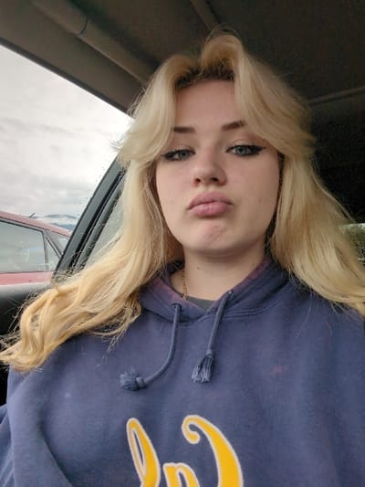 A young blonde female is depicted from the chest up, seated inside a vehicle. She has fair skin, blue eyes accentuated with black winged eyeliner, and light pink lips pursed into a pout. Her long, wavy blonde hair is styled with face-framing layers and volume at the crown. She wears a navy blue hooded sweatshirt with a partially visible yellow and white graphic at the bottom, a grey undershirt, and a thin gold chain necklace. Faint reddish-purple discoloration is present on the hoodie around the neckline. Through the vehicle's window on the left, an overcast grey sky and distant snow-capped mountains are visible, consistent with the mountainous geography of Area D (Elaho/Garibaldi), Canada. The roof of a red vehicle is partially visible outside, and raindrops or condensation are present on the window glass. The subject looks directly towards the camera.