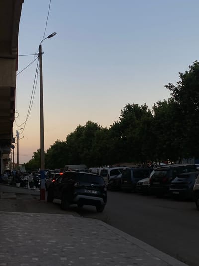 A street scene in El Hajeb, Morocco, at dusk. The sky is a gradient of light blue to pale orange. A tall, slender lamppost with wires attached stands on the left side of the image, near a building with a light-colored facade. Several cars are parked along the street, including a dark-colored SUV in the foreground on the right. Trees with dense green foliage line the right side of the street, and some people are visible seated at tables in the lower-left portion of the image, suggesting an outdoor seating area for a cafe or restaurant. The pavement in the foreground is textured and grayish.