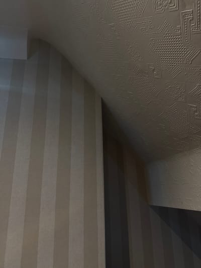 An angled wall and ceiling converge, showcasing different textures. The ceiling, adorned with intricate patterns, curves down to meet a wall covered in horizontal stripes of varying shades. Above the striped wall, a darker wall section is visible. The left bottom corner reveals a glimpse of a white architectural element, possibly a column.
