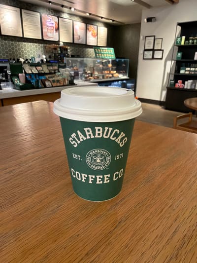 A single Starbucks coffee cup, green with white branding ("STARBUCKS EST. 1971 FRESH ROASTED COFFEE CO." and the mermaid logo), topped with a white plastic lid, rests centrally on a light brown wooden table. The table displays a distinct wood grain pattern. In the background, the interior of a Starbucks coffee shop is visible, featuring a counter with dark green hexagonal tiling. Above the counter are several menu boards listing "COFFEEHOUSE CLASSICS" and other items. Espresso machines, beverage preparation equipment, shelves with packaged food items, and a glass display case containing baked goods are discernible on and behind the counter. To the right, a wall with multiple framed pictures or certificates is present, alongside a dark shelving unit displaying various Starbucks merchandise, including mugs and coffee bags. Part of a round wooden table and a wooden chair are also visible. No individuals are present in the image. The environment is consistently lit by interior overhead lighting. While the image depicts a Starbucks interior typical of locations found across the United States, there are no specific identifying markers within the frame to confirm a precise address within Dover, United States of America.