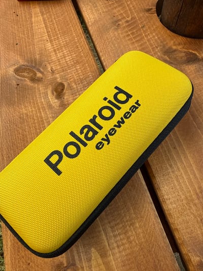 A bright yellow case for eyewear with "Polaroid" and "eyewear" printed in black is lying on a rustic wooden table. The table has a visible wood grain and appears to be outdoors, with hints of greenery and a wooden post visible in the background. The image is taken from a high angle looking down at the case. There are no people or animals present in the image. The scene is likely from Zgorigrad, Bulgaria, given the context provided, but there are no specific landmarks visible that confirm this location.