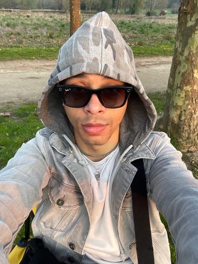 The image depicts an adult male in an outdoor setting in Plaisir, France. The subject is positioned for a close-up photograph, gazing directly towards the camera. He wears a gray camouflage-patterned hoodie with the hood up, black Ray-Ban sunglasses, a gray denim jacket, and a white t-shirt. A silver chain necklace with an intricate silver pendant is visible around his neck, and a small silver hoop earring is present in his right earlobe. A black strap, consistent with a shoulder bag, is visible over his right shoulder. A yellow object, possibly a folding chair or bag, is partially visible on the lower left. The background features a dirt path, an expanse of green grass with sparse vegetation, and several deciduous trees with bare branches. A large tree trunk with textured bark is prominent on the right side of the frame. The scene is illuminated by warm, low-angle sunlight, highlighting the subject's right side and the tree trunk.