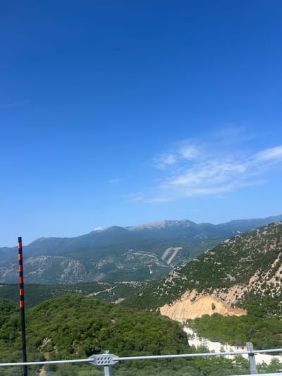 The image captures a vast mountain landscape under a clear blue sky with scattered white clouds. In the foreground, a black pole with alternating red and yellow bands is visible on the left side, alongside a wire fence. The mountains are covered in lush green vegetation, with some areas showing exposed rock and soil, particularly on the right side where a landslide appears to have occurred. The distant peaks are tinged with a lighter shade, suggesting haze or distance. The scene is indicative of a natural, undeveloped area, possibly a mountain road or viewpoint in the vicinity of Νέος Γοργόμυλος, Greece, given the context. There are no people or animals visible in the image.