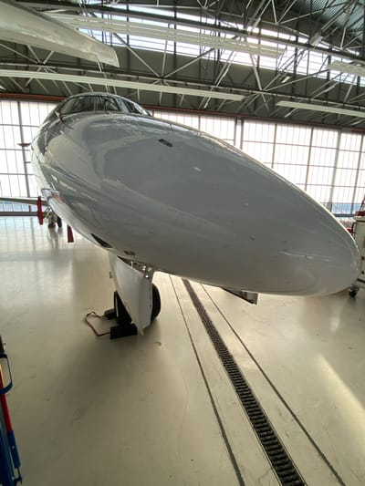 The media file displays a large white aircraft inside what appears to be an aircraft hangar in Skulte, Latvia. The primary subject is the sleek, white fuselage of an aircraft, seen from a low angle, giving prominence to its underside and nose section. The aircraft's surface is generally clean but shows some minor smudges and reflections of the hangar environment. Part of an aircraft wing, possibly with a winglet, is visible on the left side, extending upwards.

The setting is an industrial hangar with a high ceiling supported by an elaborate structure of metal trusses and beams. Large, gridded windows along the walls or ceiling allow ample natural light to illuminate the space, suggesting it is daytime. The concrete floor of the hangar features a long, linear drainage channel with a grated cover, running parallel to the aircraft's length.

There are no people visible in the image, implying the aircraft is either undergoing maintenance, storage, or is awaiting operations. The presence of the hangar, aircraft, and specialized equipment points to an aviation facility. A red "REMOVE BEFORE FLIGHT" tag is partially visible near the aircraft's lower fuselage, attached to a component, indicating that the aircraft is prepared for or undergoing procedures that require such safety warnings.