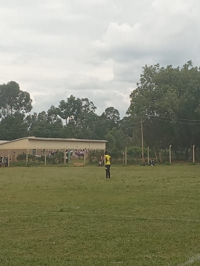 The image depicts an open, grassy field under an overcast sky in Chevaywa ward, Kenya. A single individual, dressed in a yellow long-sleeved shirt and dark pants, stands in the mid-ground with their back to the viewer, facing towards a cluster of structures in the background. To the left, a light-colored, single-story building with a dark roof and multiple windows is visible. In front of this building, a clothesline with various garments hangs. A fence constructed from wooden posts and wire runs horizontally across the mid-background, extending from the left past the building and into dense foliage on the right. Several other individuals are discernible near the fence and building, some seated or standing. Lush green trees form a dense line along the horizon behind the structures. The ground is covered with natural green grass, appearing somewhat uneven.