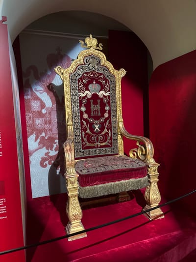 An ornate, gilded throne chair upholstered in crimson velvet and intricate gold embroidery is displayed in a museum setting, likely within the Grand Palace of Gatchina, Russia. The chair's backrest features a crown emblem and detailed patterns, while the seat and arms are also heavily ornamented. The throne is placed on a raised red carpeted platform. Behind the chair, a large wall display showcases a red and white pattern of what appears to be a double-headed eagle crest, a national symbol of Russia, with smaller crests visible within its wings. The surrounding walls are painted a deep red, and an arched ceiling is visible above. A black stanchion cord runs horizontally in the foreground, indicating a display area that is cordoned off from visitors. The lighting casts shadows on the throne and the backdrop, highlighting the richness and historical significance of the artifact.