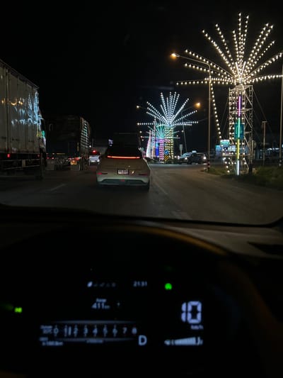The image captures a nighttime scene on a road in Lang Suan, Thailand, likely during a festival or event given the elaborate light displays. The primary focus on the left side is a large, starburst-shaped light decoration made of numerous small bulbs. Another similar, smaller light decoration is visible below it. These lights illuminate the dark surroundings, creating a festive atmosphere.

The scene is viewed from inside a vehicle, as indicated by the dashboard elements on the right side of the image. The dashboard displays digital readouts including a gear indicator showing "D" and a series of numbers, likely representing speed or trip information. A green indicator light is also visible.

The road appears to be a multi-lane street, with several vehicles, including trucks and cars, visible. Some vehicles have their brake lights on, suggesting they are stopped or moving slowly. The presence of traffic and the illuminated decorations suggest a populated area, possibly a town center or a roadside attraction. The time of day is clearly night, and the weather appears to be clear, allowing for the vibrant display of lights.

Text is visible on a license plate of a white car: "บฉ 0225". Other signs with text are present along the road, including traffic signs with arrows and some in what appears to be Thai script. The overall impression is one of a lively, illuminated street at night, possibly a market or fairground, viewed from the perspective of a driver.
