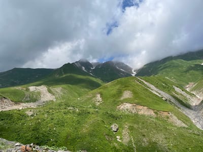 This is a landscape photograph of rolling green mountains under a cloudy sky. The mountains are covered in lush green grass, with patches of bare earth and rock visible in some areas. There are also streaks of snow on the higher peaks. The sky is filled with dramatic, dark clouds that are beginning to break, allowing some sunlight to filter through. The overall impression is one of a rugged and wild natural environment. The location is identified as Almasiani, Georgia.