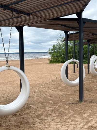 The image depicts a beach scene in Sestroretsk, Russia, featuring an outdoor seating area with a slatted wooden roof supported by black metal pillars. Several white, circular swings, resembling large donuts, hang from chains suspended from the roof structure. The swings are positioned over a sandy beach, with the Gulf of Finland visible in the background. The water is choppy, and the sky is overcast. There are a few distant figures walking on the beach, and some greenery, likely trees and bushes, are present behind the beach. The sand shows some footprints and traces of tire tracks. The overall atmosphere is serene and suggests a public recreational space.