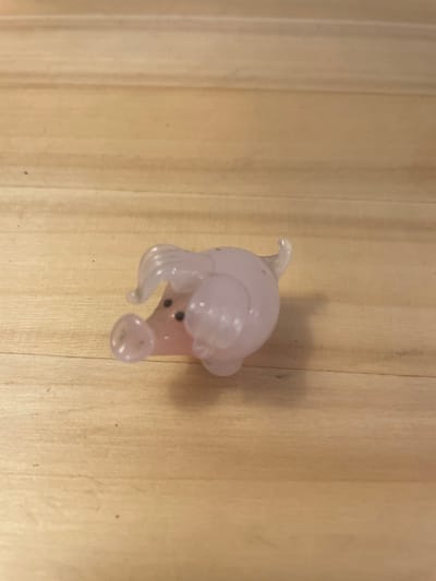 A small, translucent pink glass figurine of a pig is positioned on a light-colored wooden surface. The pig is lying on its side, facing towards the left of the frame. Its snout is prominent, and two small black dots represent its eyes. The figurine has a curly tail and ears that are characteristic of a pig. The wooden surface appears to be made of planks with visible grain. The background consists of more of the wooden surface. The lighting is soft, casting subtle shadows from the figurine. There are no people or any other objects visible in the image. The scene is static and does not depict any action. The location is unknown.