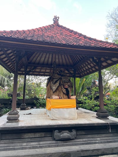 The image shows a Balinese Barong mask and costume displayed on a raised platform under a tiled pavilion in Dawan, Indonesia. The Barong, a mythical creature from Balinese folklore, is adorned with elaborate headdresses and a shaggy, straw-like body. It is seated on a white cushion, draped with a bright orange cloth. The pavilion features dark wooden beams and a red-tiled roof topped with a decorative finial. The platform is covered with a woven mat. In the background, lush green foliage, trees, and stone structures typical of a Balinese temple complex are visible. The sky is overcast. There are no people present in the scene.