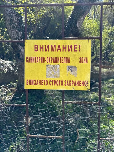 A yellow sign with red lettering reading "Внимание!" (Attention!) and "Санитарно-охранителна зона" (Sanitary-security zone) is posted on a metal fence. The sign also says, "влизането строго забранено" (entry strictly forbidden). It is partially obscured by the rusted metal fence and some vegetation. The sign is located in the town of Zemen, Bulgaria, at the coordinates 42.467, 22.738. The area surrounding the sign is forested and overgrown with vegetation. The sign appears to be in a natural or outdoor setting.