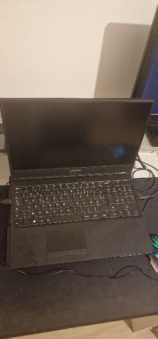 The scene depicts a workstation located in Blaye, France. A black Legion brand laptop computer, closed or with a dark display, is positioned centrally on a large, dark-colored mousepad. The laptop's keyboard features a French AZERTY layout, with a USB-A cable connected to its left side. To the right of the laptop, a coiled blue cable and a partially visible peripheral with green and purple highlights are present on the mousepad. A black cable runs horizontally across the foreground of the mousepad. In the background, a light-colored wall is visible, along with the dark screen of an upright monitor casting a shadow on the wall to the right. No individuals or active interactions are observed in the image.