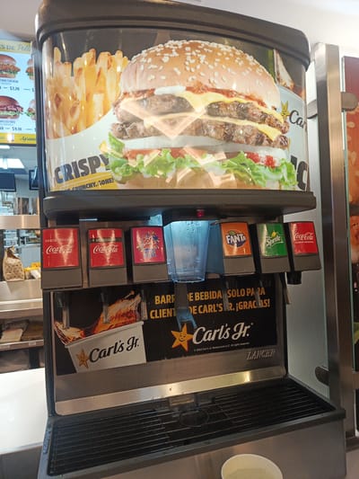 The image shows a self-service soda fountain machine at a Carl's Jr. restaurant. The machine has six beverage options: Coca-Cola (original and sugar-free), Fanta Fresa, Fanta Orange, Sprite, and Coca-Cola (original). Above the dispensers is a large advertisement featuring a burger and fries. The machine is marked with the Carl's Jr. logo and the phrase "BARRA DE BEBIDA SOLO PARA CLIENTES DE CARL'S JR. ¡GRACIAS!". Below the dispensers is a drip tray. A small cup is visible at the bottom of the image. The restaurant setting is discernible in the background, with a menu board and other food service equipment visible. Based on the phrase "BARRA DE BEBIDA..." which means 'Drink Bar', the location is likely Ecuador. Given the American fast food chain being present, the city Samborondón, Ecuador is a possible location.