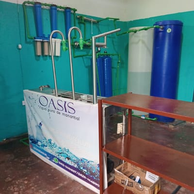 The image shows an Oasis water purification system, identifiable by the branded signage "OASIS 'Agua pura de manantial'" and contact details. The system includes blue cylindrical tanks connected by green pipes, suggesting a multi-stage filtration process. Stainless steel support structures are also visible. The scene's location may be Duaca, Venezuela, given the provided phone numbers and product availability. The room is painted light green/teal. A small brown cardboard box is placed on a shelf.