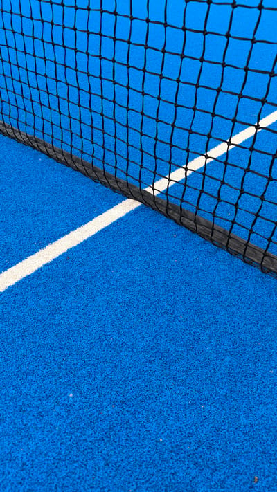 The image shows a close-up view of a blue court with a black net in the foreground. A white line runs diagonally across the court, partially obscured by the net. The court surface appears to be textured. The scene is static and lacks any human presence or activity. The image is likely taken at an outdoor sports facility in Turgoyak, Russia, though there are no specific landmarks or identifying features to confirm this location. The lighting suggests it is daytime.