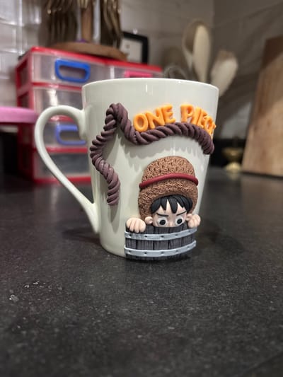 This image captures a close-up, eye-level view of a custom-decorated white ceramic mug resting on a dark, textured countertop in Mu'adammiyat ash-Sham, Syria. The mug is adorned with polymer clay embellishments depicting characters and elements from the "One Piece" manga/anime series. Orange raised letters on the mug spell "ONE PIECE," positioned above a dark brown braided rope design. Below this, a stylized character resembling Monkey D. Luffy, wearing a brown straw hat with a red band, is depicted peeking over the top edge of a grey barrel-like structure. The character has black hair and wide, expressive eyes. The mug's handle is visible on the left side. The background, largely out of focus, shows red and clear plastic storage drawers stacked vertically on the left, and various kitchen utensils, possibly wooden or metallic, in a container on the right, alongside what appears to be a wooden cutting board. The surface beneath the mug is a dark grey or black countertop with subtle speckling and texture.