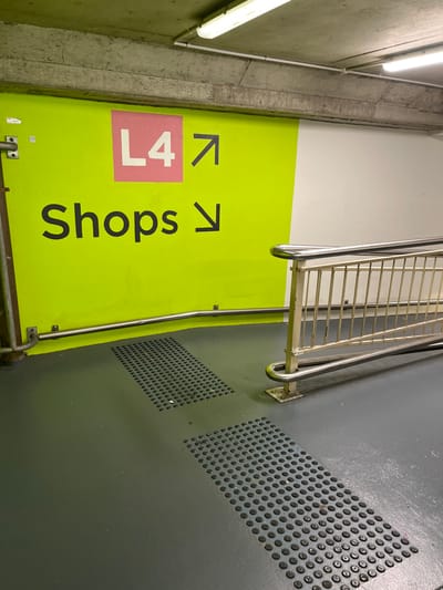 This image captures an indoor area, likely a walkway or stairwell transition within a commercial building such as a shopping center in Indooroopilly, Australia. No people are visible, and there is no specific activity taking place, rather the scene depicts functional architecture designed for navigation.

The prominent feature is a bright lime green wall panel that stretches across the middle of the frame. On this panel, clear directional signage is displayed: the text "L4" is enclosed within a pink square, accompanied by an arrow pointing upwards and slightly to the right. Next to it, the word "Shops" is written in large black letters, with an arrow pointing to the right. These signs indicate directions to a specific level (Level 4) and to retail shops.

To the left of the green wall, a rugged concrete wall is visible, featuring two horizontal fluorescent tube lights providing artificial illumination. Conduits or pipes run along this concrete surface. To the right of the green wall and railing, there is a dark grey floor, possibly made of concrete or a similar industrial material. This floor has two distinct rectangular sections of perforated metal, resembling grates or textured tactile paving. A sturdy metal railing, appearing to be a handrail for stairs or a ramp, runs along the base of the green wall and extends upwards. Another section of railing is visible higher up, suggesting multiple levels or a longer pathway.

The lighting appears artificial, suggesting an indoor setting regardless of the time of day or weather outside. The overall impression is one of a utilitarian yet clearly marked interior space, guiding visitors through a multi-level complex.