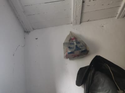 The image depicts an interior corner of a room, possibly a storage area or unfinished space, in Ivanovka, Kyrgyzstan. The ceiling is constructed of rough, white-painted wooden planks and beams. The walls are white and show signs of wear, including cracks and minor imperfections.
Hanging from a hook on the wall is a transparent plastic bag containing various items, appearing to be refuse or discarded goods. Some of the items within the bag have red and blue labels. In the lower right portion of the image, a dark-colored object, possibly a bag or piece of clothing, is partially visible, with a translucent plastic material inside it. The overall impression is of a utilitarian and somewhat neglected space.