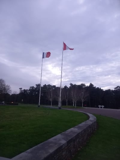 Two flags fly beneath a cloudy sky. One is red and white, possibly the flag of Austria, the other is red, white, and blue. In the background, trees line the horizon. To the right, a concrete wall curves alongside green grass. The overall setting appears to be a park or outdoor memorial site.