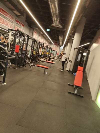 A gym's interior features exercise equipment like weight machines and benches with red cushions. People are working out, and overhead, industrial-style ventilation ducts run along the ceiling, illuminated by linear lights. Motivational signage adorns the walls, and the space is open and well-lit.