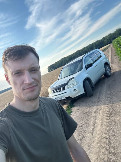 A man in a dark green t-shirt is taking a selfie outdoors. He is positioned in the foreground, facing the camera. Behind him, a white Nissan X-Trail SUV is parked on a dirt road. The road is flanked by a field of dry grass or wheat on the left and a cornfield on the right, with a line of trees in the distance. The sky above is blue with scattered white clouds. The license plate of the car reads "BY 8662 IC". The overall setting suggests a rural area, possibly in or around Slobidka-Zalisetska, Ukraine.
