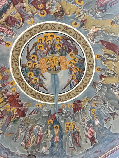 This image is a detailed fresco on a domed ceiling within a religious building in Nepraznentsi, Bulgaria. The central composition features a large circular medallion depicting numerous haloed figures, predominantly winged angels, arranged in concentric rows on a light blue background. Many angels hold rectangular, gold-paneled objects or books. A stylized, light blue architectural element extends downwards from the center. This medallion is encircled by a broad, dark brown border patterned with a lighter tan floral or vine motif.
Surrounding the central medallion is a sprawling scene set against a grey-blue sky and a landscape of stylized, jagged rock formations in shades of brown and grey. This outer scene depicts numerous haloed human figures and angels. In the lower sections, a distinct area of dark blue water contains several naked or partially clothed figures in distress, some being pulled from the water by winged angels. Small, red fish are visible in the water. Elsewhere in the outer scene, figures are shown emerging from rocky tombs, ascending or descending steep terrain, and interacting with each other. Some figures carry objects, such as rectangular boxes or scrolls, while others are depicted pulling a two-wheeled cart drawn by a brown ox. The fresco utilizes a color palette dominated by blues, greys, browns, reds, and golden yellows for halos.