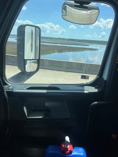 The image is taken from the interior of a vehicle, looking out through the driver's side window and a portion of the windshield. The vehicle appears to be a large truck, as indicated by the size of the side mirror and the interior components. Outside, a bright, sunny sky with scattered clouds is visible. The vehicle is on a bridge or elevated road, with a concrete barrier running along the edge. Beyond the barrier, a body of water, likely a river or bay, is visible, with marshy vegetation along its banks and in the distance. The water reflects the sky. On the windshield, near the bottom right corner, a small American flag sticker is affixed. Inside the vehicle, in the lower part of the frame, a blue plastic container with a red and white spigot is visible. The interior of the truck is dark, with shadows obscuring most details. The location is identified as Riceboro, United States.