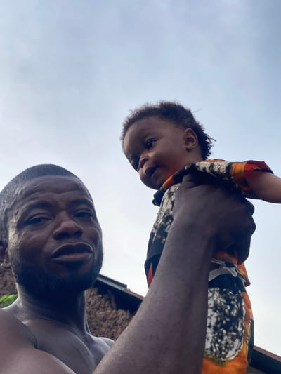 An outdoor, low-angle, close-up, chest-up, selfie-style portrait depicts a man and a toddler against a cloudy sky. The man, on the left, faces the camera with his head tilted slightly. His eyes are open, and he appears to be speaking. He has short, dark hair, a beard, and is shirtless. The toddler, held by the man's right arm and shoulder, is turned to the toddler's left and looking away from the camera. The toddler has curly hair and is wearing a colorful, patterned shirt. In the background, a thatched roof structure with wooden beams is visible. The overall lighting is soft and diffused, suggesting an overcast day. The image is likely taken in Unwana, Nigeria, given the visual cues of the background structure.