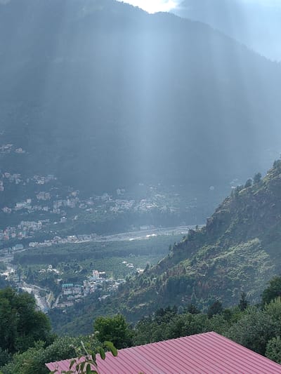 The image depicts a valley view in Siumsa, India, characterized by a hazy atmosphere with visible sunbeams filtering through the mist. The foreground features a red corrugated metal roof and lush green trees. The midground reveals a densely populated town nestled within a valley, with numerous buildings of varying sizes and colors. A river or stream is visible flowing through the town, and it is surrounded by extensive green vegetation and forests that climb the surrounding hillsides. The background is dominated by the imposing, mist-covered mountains, with a hint of the sky visible at the very top. The overall impression is one of a picturesque and serene mountain landscape.