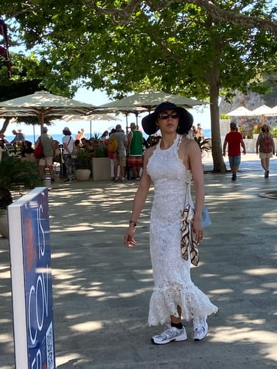 The image depicts a woman in a white lace dress and a wide-brimmed hat walking on a paved walkway in Dubrovnik, Croatia. She is wearing sunglasses and carrying a small light blue purse. In the background, there are several people, outdoor umbrellas, trees, and the sea. A sign with blue and white lettering is visible in the foreground on the left. The scene appears to be a sunny day, with shadows cast by trees on the ground.