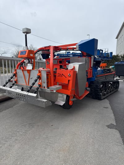 The image shows a piece of heavy machinery or equipment, possibly agricultural or industrial, located outdoors on what appears to be a paved road or parking area. The color scheme of the machine is primarily orange, silver, and blue. It features complex mechanical structures, including tracks and what seem to be sensor systems or control panels. The equipment is standing near a building or structure, which indicates that it could be in an industrial area. Based on the title, we can place the machinery in Logatec, Slovenia.
