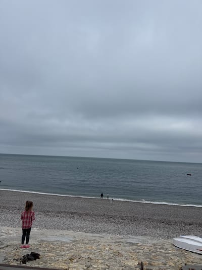 This image shows a wide, overcast view of the beach and sea in Étretat, France. In the foreground, a young girl with blonde pigtails, wearing a red and white plaid shirt and dark pants, stands facing away from the camera, looking out at the water. She has pink shoes on. To her left, on the rocky ground, are some dark, possibly metal, objects. The beach is covered in pebbles. In the distance, along the shoreline, two figures can be seen walking, and a third figure appears to be standing near the water's edge. A small boat is visible on the water, to the right of the frame. The sea is a dark, deep blue, and the sky is filled with thick, grey clouds. The overall impression is of a somber, cloudy day by the coast.