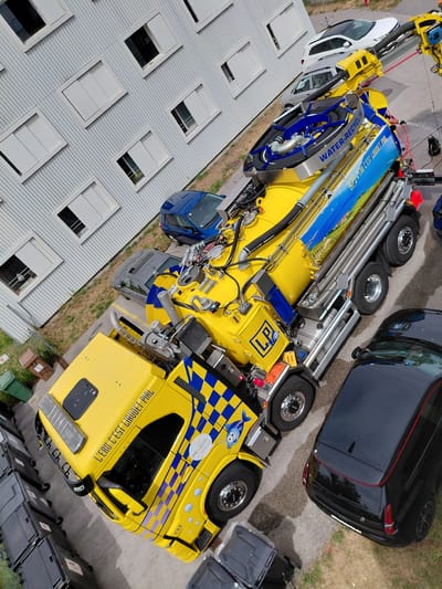 The image shows a yellow and blue vacuum truck parked in front of a corrugated metal building with white-framed windows. The truck has a large tank and various hoses and equipment. A black car is parked to the right of the truck, and a silver car and a blue hatchback are parked behind it. The scene appears to be in an industrial or commercial area, possibly a parking lot. The building's design and the presence of several vehicles suggest an urban or suburban setting. The overall impression is a still-life scene, with no humans or animals visible and no overt actions taking place. The location of Ecublens, Switzerland is not evident from the visual information alone, but the image is consistent with a location in that country.