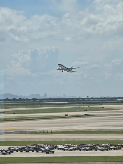 An airplane is ascending into a partially cloudy sky over an airport. The airplane, predominantly white with blue accents, is captured mid-flight, its landing gear retracted. In the background, a distant city skyline is visible, obscured by haze and featuring numerous tall buildings. The airport itself features multiple runways and taxiways, with a long row of luggage carts lined up in the foreground on the tarmac. The scene suggests an operational airport environment, likely in a densely populated urban area, consistent with the location of Bao'an District, China.