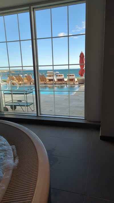 The image depicts a view from indoors, looking through a large window with a grid of panes, out towards a body of water. In the foreground, a portion of a circular, ribbed, tan-colored structure, likely part of a hot tub, is visible on the left. The floor inside appears to be dark tile.
Through the window, a swimming pool with clear blue water is in the mid-ground. Around the pool, several wooden lounge chairs are arranged. To the right of the pool, a red umbrella is closed and upright, with a small, round, textured ottoman nearby. Beyond the pool and lounge chairs, a body of water, possibly the sea, stretches to the horizon under a bright blue sky with scattered white clouds. The coastline in the distance appears rocky and somewhat barren. The scene is likely a resort or hotel with outdoor amenities.
The location is identified as Larkollen, Norway. This suggests the view is of the Oslofjord or the Skagerrak. The outdoor setting with a pool and lounge chairs implies a recreational or resort area within Larkollen. The presence of the hot tub in the foreground indicates an indoor spa or relaxation area connected to the outdoor view.